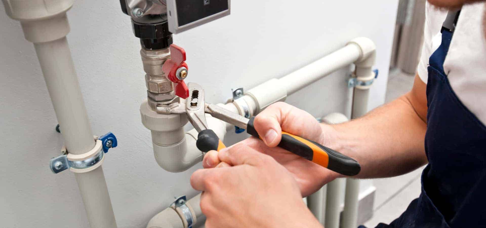 A person is using pliers to adjust a valve on a network of white pipes mounted on a wall. They are holding the valve steady with one hand while turning it with the pliers in the other hand. The setting appears to be an indoor plumbing setup.