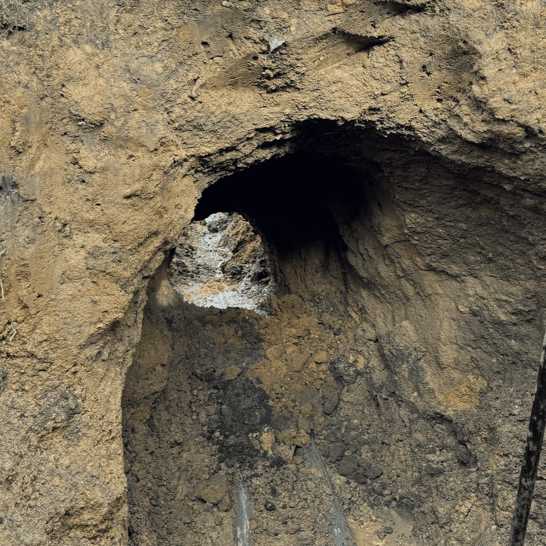 A rocky, rough-surfaced cave entrance with dim lighting resembles the aftermath of a sewer line replacement project. The uneven path, with dirt and loose stones scattered around, mirrors the jagged walls that reveal a rugged and natural appearance.