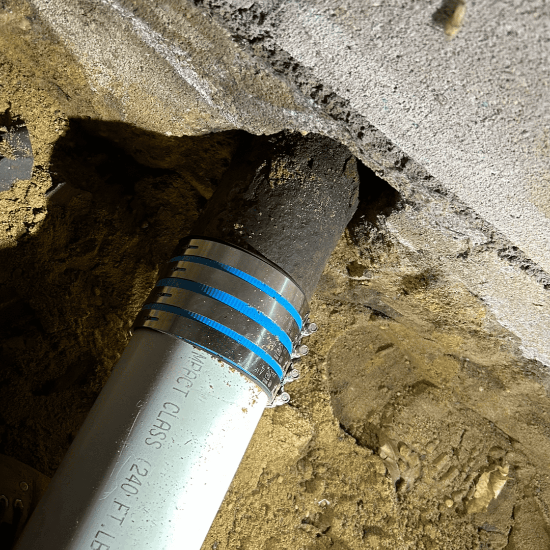 A broken underground sewer line with a blue and silver clamp is partially repaired, emerging from the sand and soil next to a concrete wall.