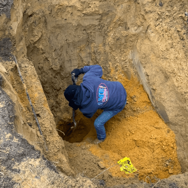 A person wearing a blue hoodie and jeans is digging a hole in the ground with a shovel, likely for sewer line replacement. The soil is brown and orange, with small yellow caution tape partially buried in the dirt, signaling ongoing work.
