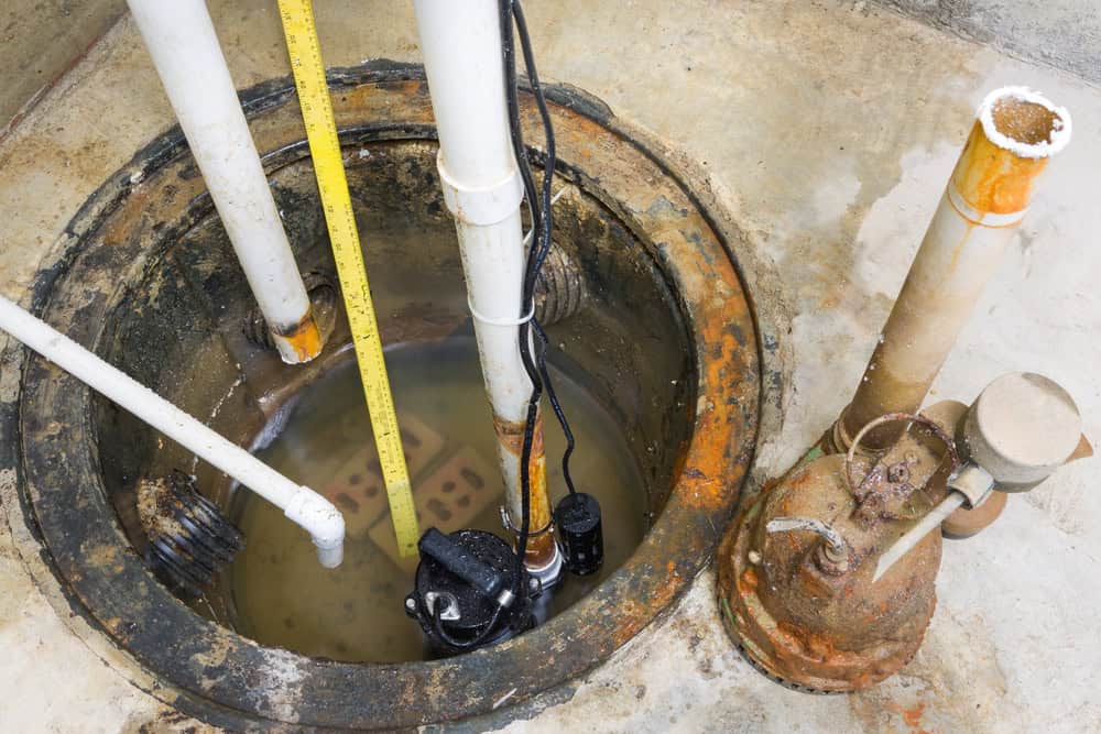 A close-up view of a sump pump in a basement, intertwined with sewer line repair pipes and standing water. The concrete floor and walls show signs of rust and wear. The setup includes a yellow measuring stick and a smaller metal pump nearby.