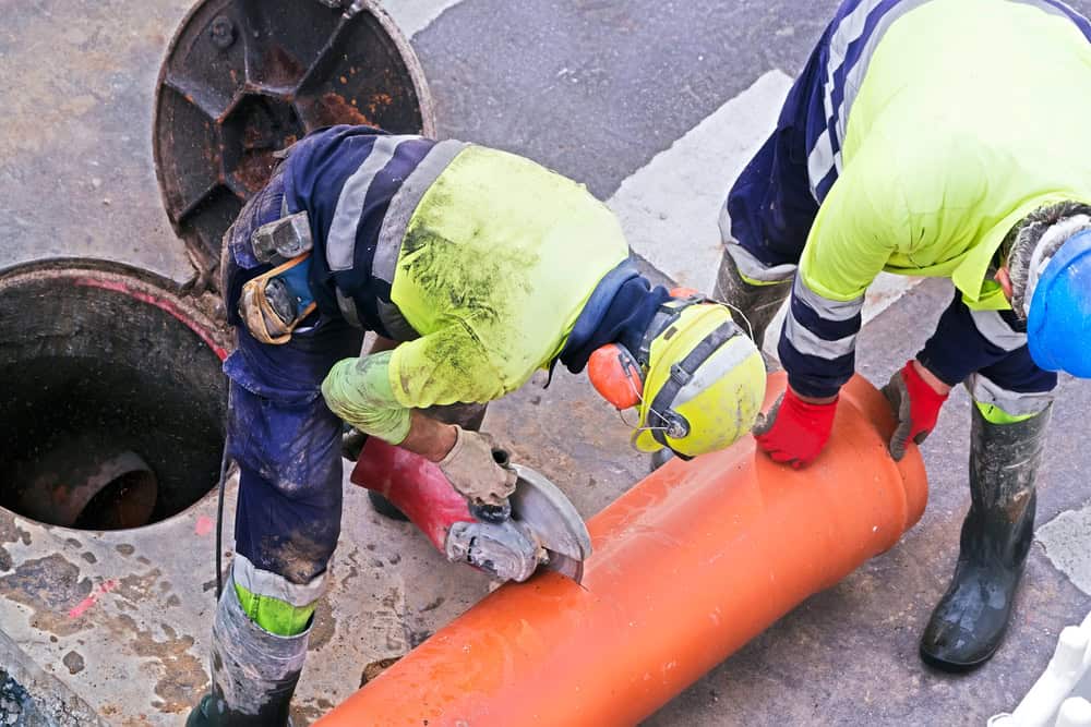 Two construction workers in high-visibility clothing and helmets are cutting an orange pipe with a power tool on a road surface near an open manhole. The scene suggests street maintenance or plumbing work.
