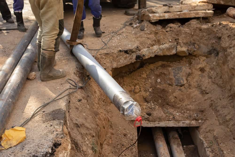 Workers in rubber boots are installing a large metal pipe into a trench. The ground is excavated, revealing existing pipes beneath. The scene is outdoors with exposed dirt and construction materials scattered around.