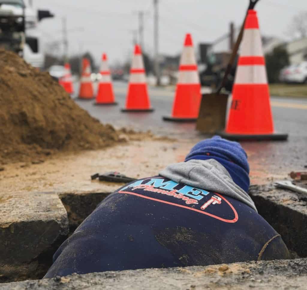 A construction worker in a blue jacket, with AME Plumbing equipment nearby, is partially visible while working in a hole on the street. Several orange traffic cones line the background, marking the work zone amid scattered tools and freshly dug dirt.