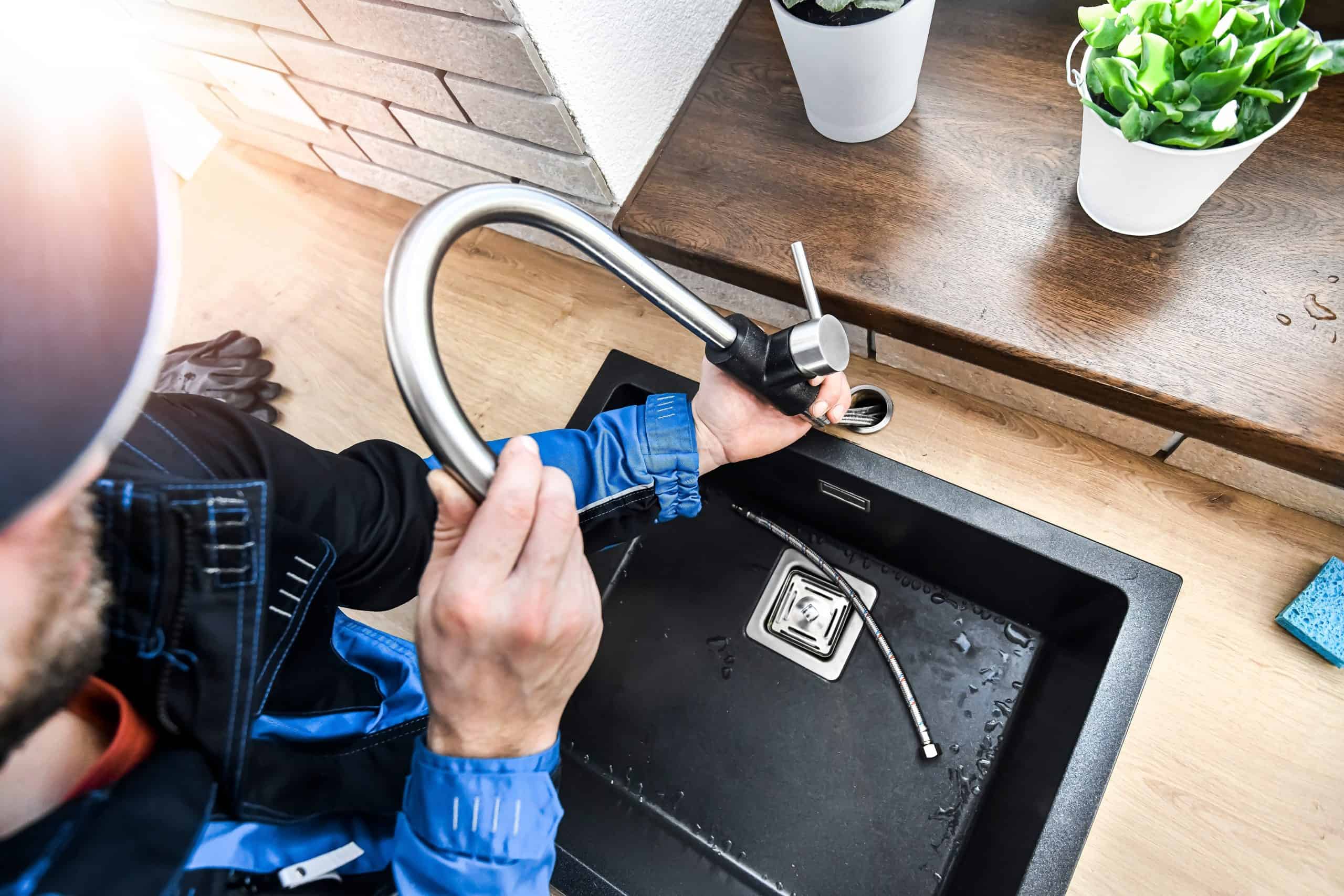 A plumber in a blue uniform installs a faucet over a black kitchen sink. A metal hose lies inside the sink. Two potted plants sit on the countertop, and sunlight streams in from the upper left corner.