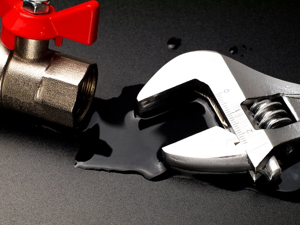 A metallic wrench lies next to a slightly open brass valve with a red handle. A small puddle of water surrounds the tool and valve on a black surface, indicating a leak.
