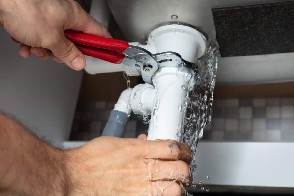 Person using red-handled pliers to fix a leaking white pipe under a sink. Water is spilling from the pipe, with droplets splashing around. The background shows a tiled wall.