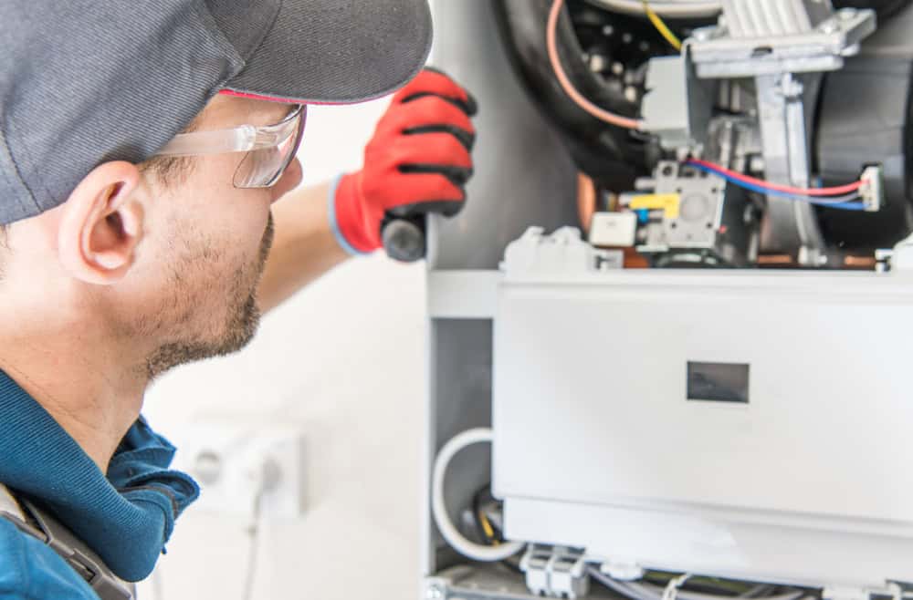 A technician wearing a cap and safety glasses is inspecting the interior of a heating or air conditioning unit. He holds a flashlight and wears a red glove on one hand. Various wires and components are visible inside the unit.