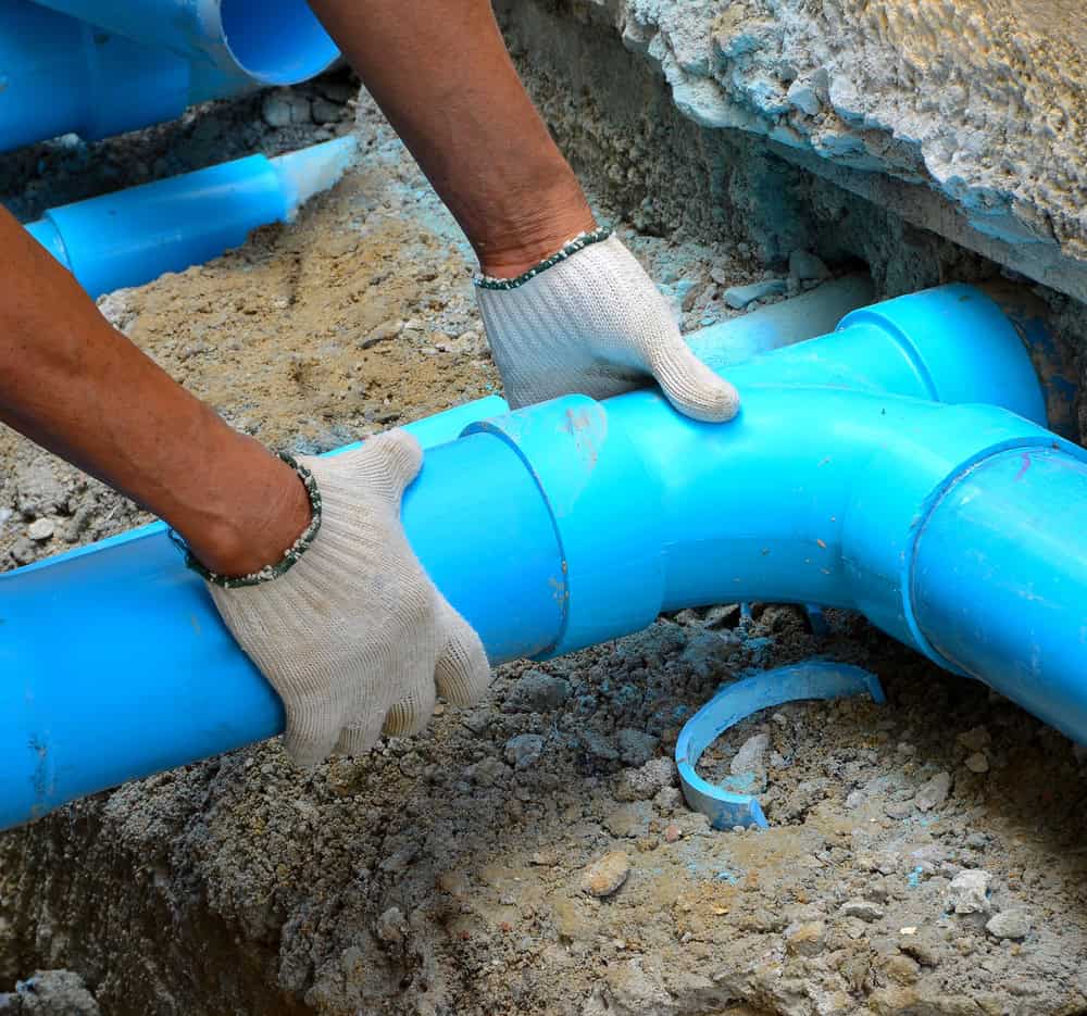 A person wearing white gloves is connecting blue PVC pipe fittings in a trench surrounded by dirt.