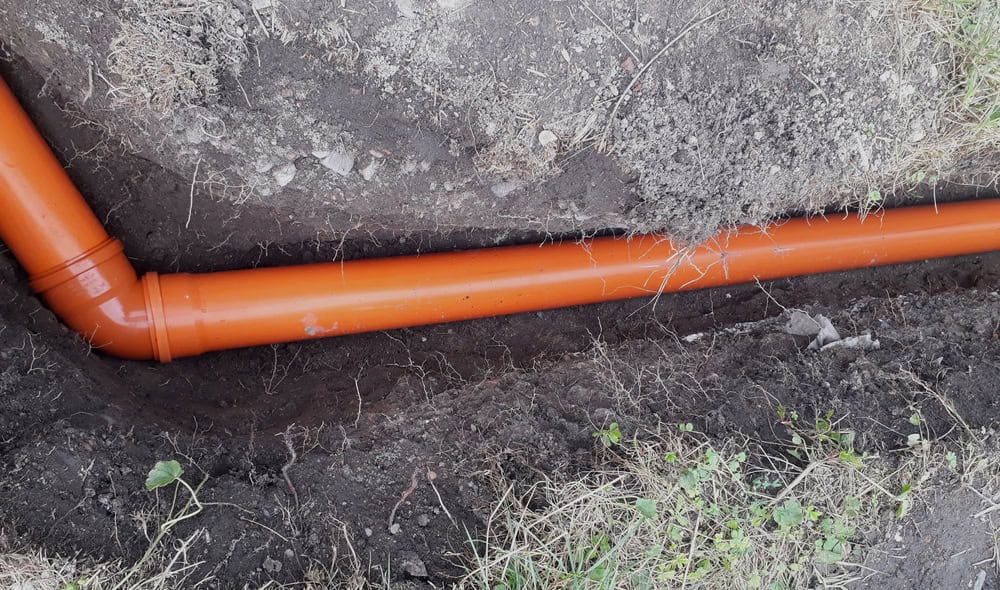 Orange drainage pipe installed underground in a shallow trench, surrounded by soil and patches of grass. The pipe features an elbow joint to redirect the flow.