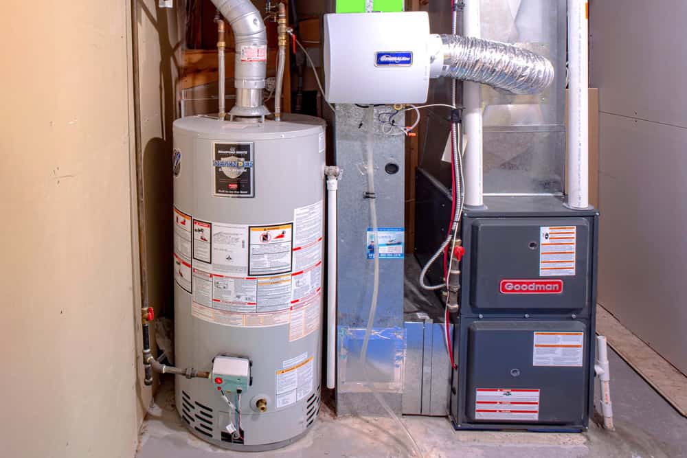 A home utility area featuring a water heater on the left and a furnace on the right. The heater has various labels and pipes, while the furnace is branded "Goodman" with ducts and a metal venting system above.
