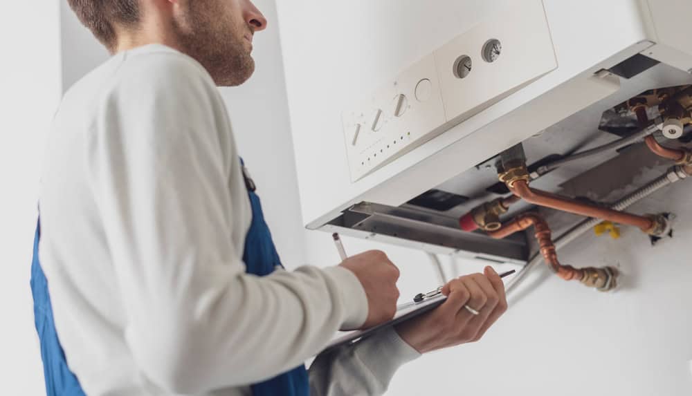 A technician in a blue overall inspects a gas boiler. They are holding a clipboard and appear to be examining the open panel, checking for maintenance or repairs. The focus is on the boiler's internal components and the technician's attention to detail.