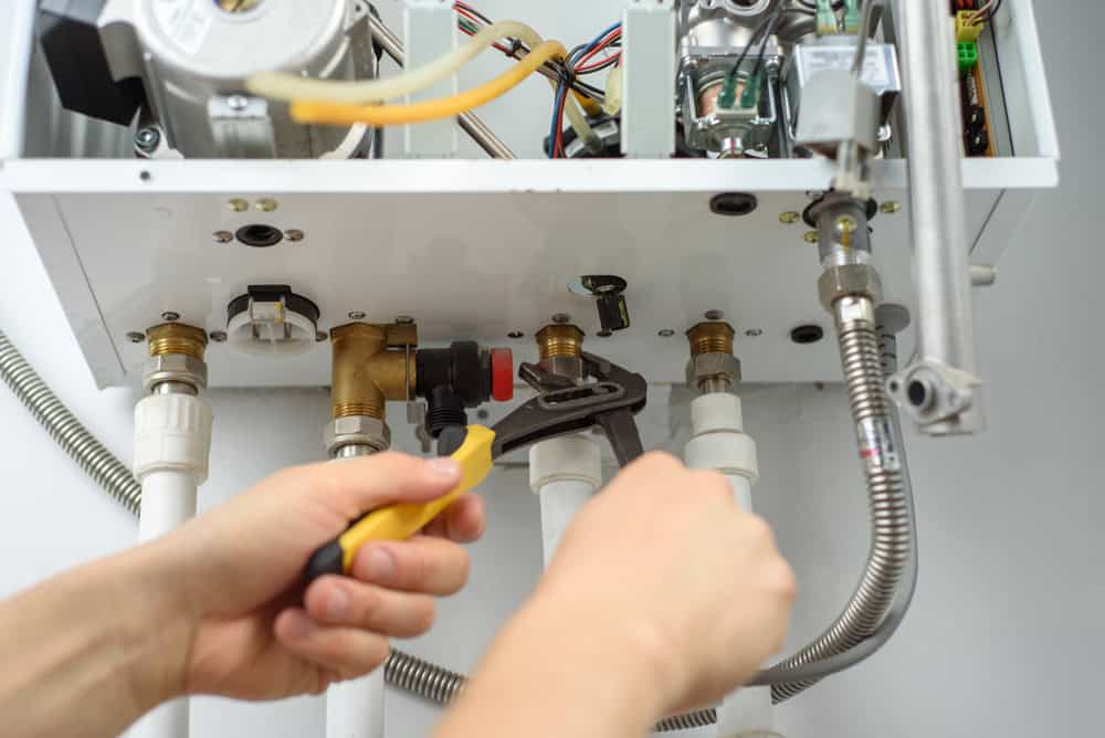 A person using a wrench to tighten a pipe connection on the underside of a boiler or water heater, with various tubes and wires visible.