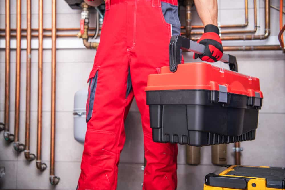 A person wearing a red jumpsuit holds a toolbox in a mechanical room. Copper pipes and equipment are visible in the background, indicating a plumbing or maintenance setting.