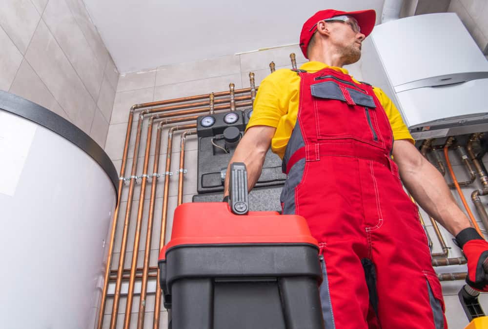 A plumber in red overalls stands in a utility room with various pipes and a water heater. He holds a tool case and looks to the side. The room has tiled walls and a modern heating system.