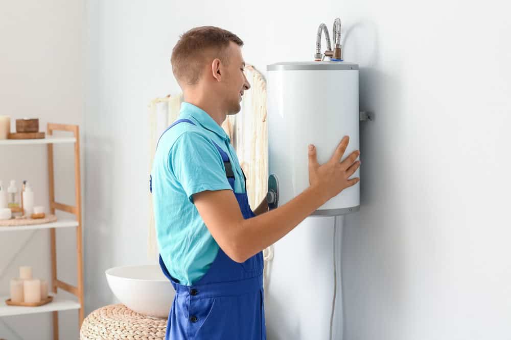 A man in blue overalls and a light blue shirt is installing or inspecting a wall-mounted water heater in a bathroom. The room is bright and contains shelves with towels and toiletries.