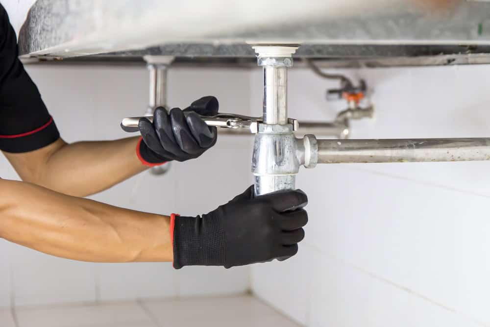 A person wearing black gloves uses a wrench to tighten a pipe under a sink. The scene shows a close-up of the plumbing work in a tiled environment.