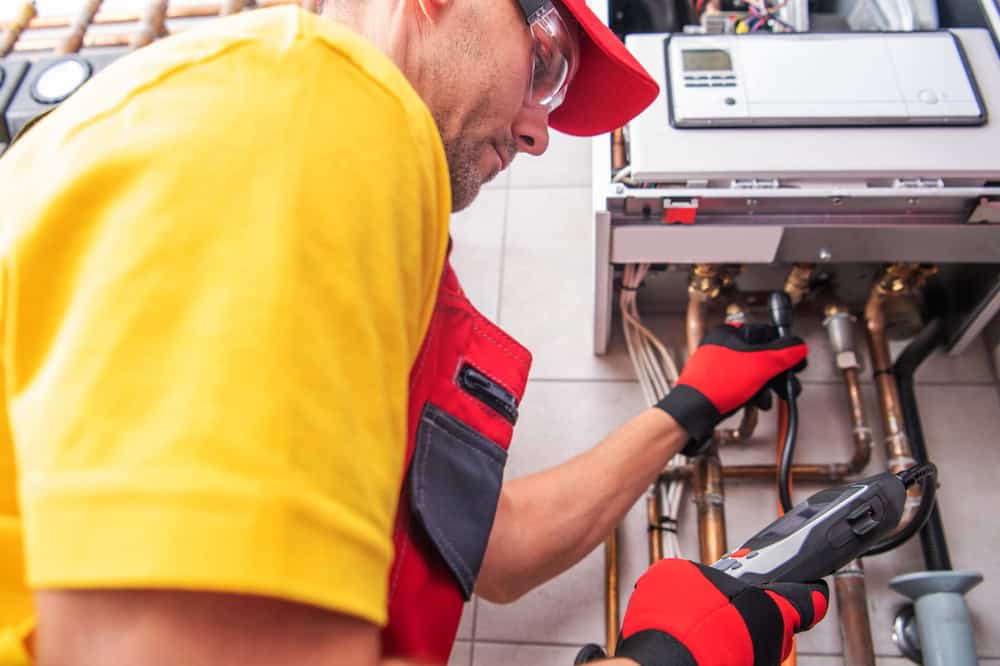 A technician wearing a red cap, yellow shirt, and red gloves is inspecting a complex network of pipes connected to a boiler or heating system. They are using a handheld device, possibly for diagnostics or maintenance.