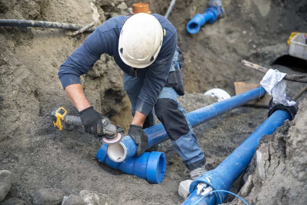 A construction worker wearing a helmet and protective gear is cutting a blue PVC pipe with a power tool in a dirt trench. Blue pipes and tools surround the worker in the excavation site.