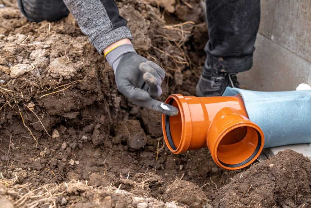 A person wearing gloves is connecting orange PVC pipes in a trench filled with soil. The pipes are part of a plumbing or drainage system. Construction materials and earth are visible around the work area.