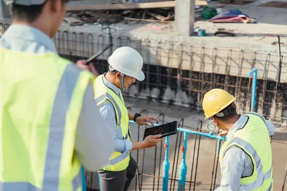 Three construction workers wearing safety vests and helmets inspect a site. One worker is using a tablet, while the others observe the structure. They are near reinforcing steel bars and concrete.