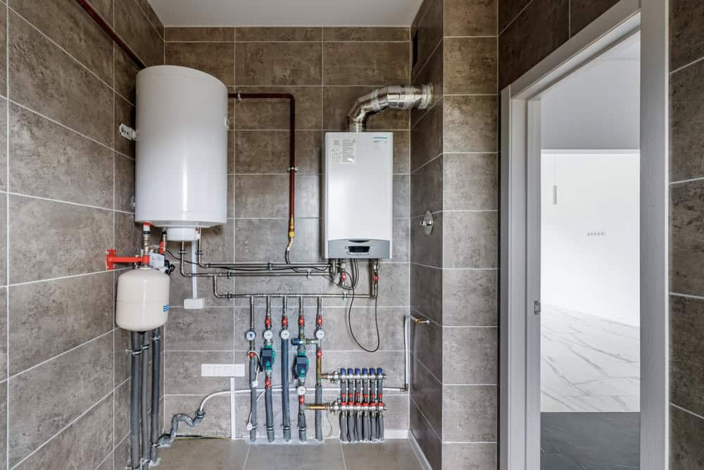 A utility room with a water heater, pipes, and a boiler attached to a tiled wall. The room has a tiled floor and an open doorway leading to a bright adjacent space.