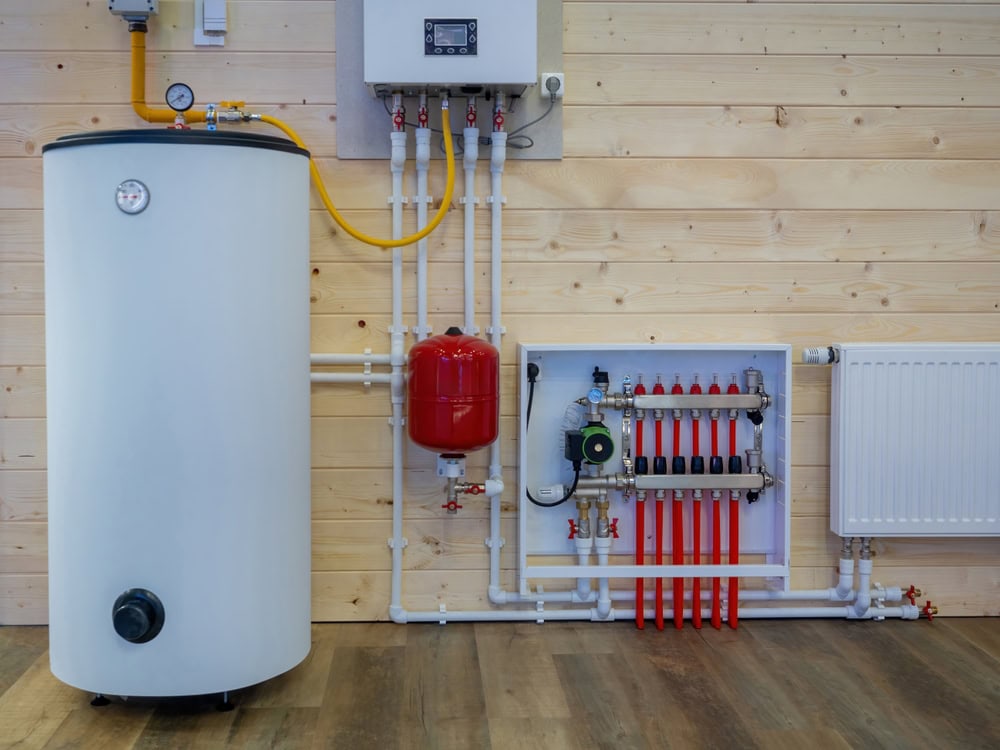 A heating system setup against a wooden wall includes a large white water tank with attached pipes, a red expansion tank, a manifold with red valve controls, and a radiator. The setup is interconnected with various pipes and valves.