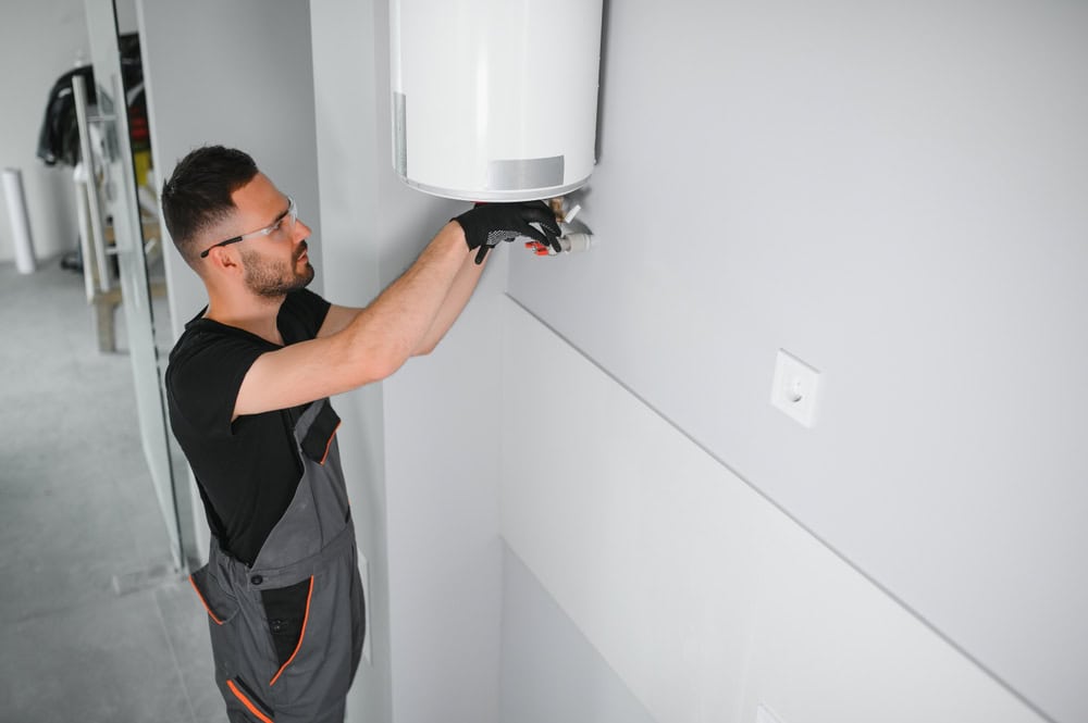 A technician wearing glasses and gloves is installing or repairing a white wall-mounted boiler in a modern room. He is focused on his task, and the room has light gray walls and minimalistic decor.