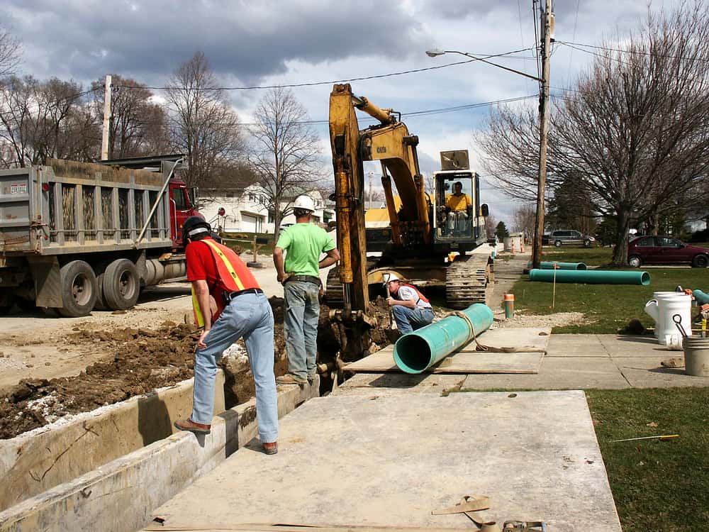 Construction workers installing large green pipes in a residential neighborhood. One worker stands on a concrete slab while others guide a pipe into a trench. An excavator and a dump truck are visible. Trees and houses line the street.