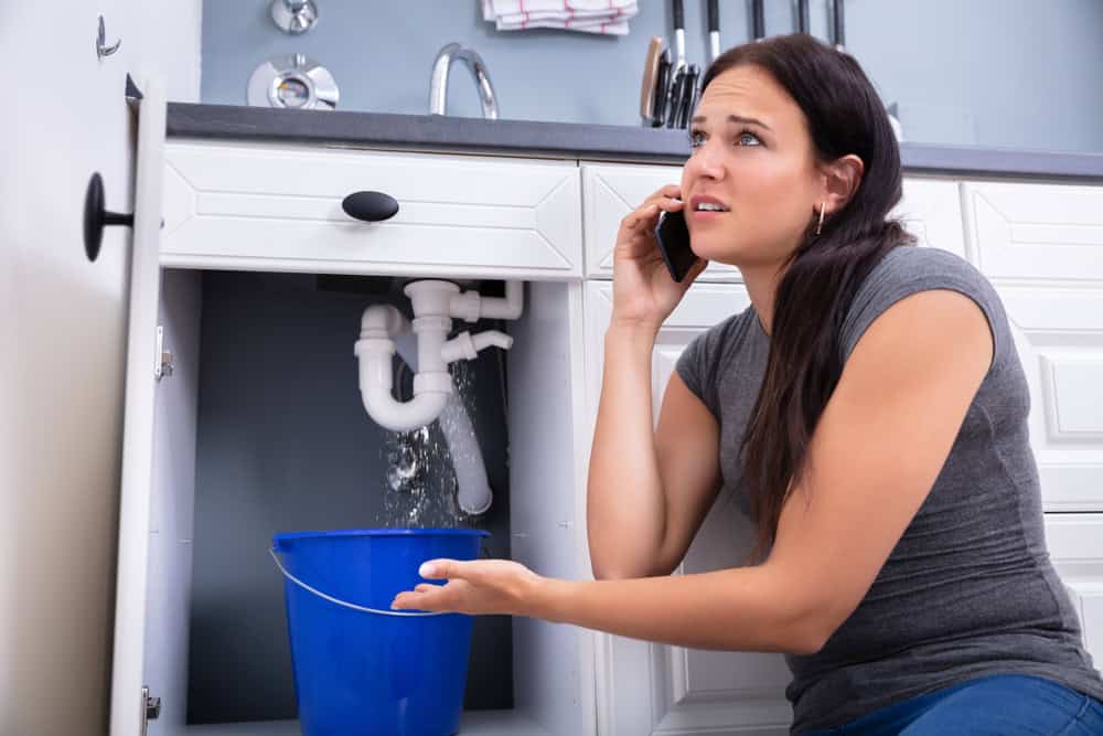 A frustrated woman is on the phone, kneeling in front of a kitchen sink cabinet with a leaking pipe. Water is pouring into a blue bucket placed below the pipe.