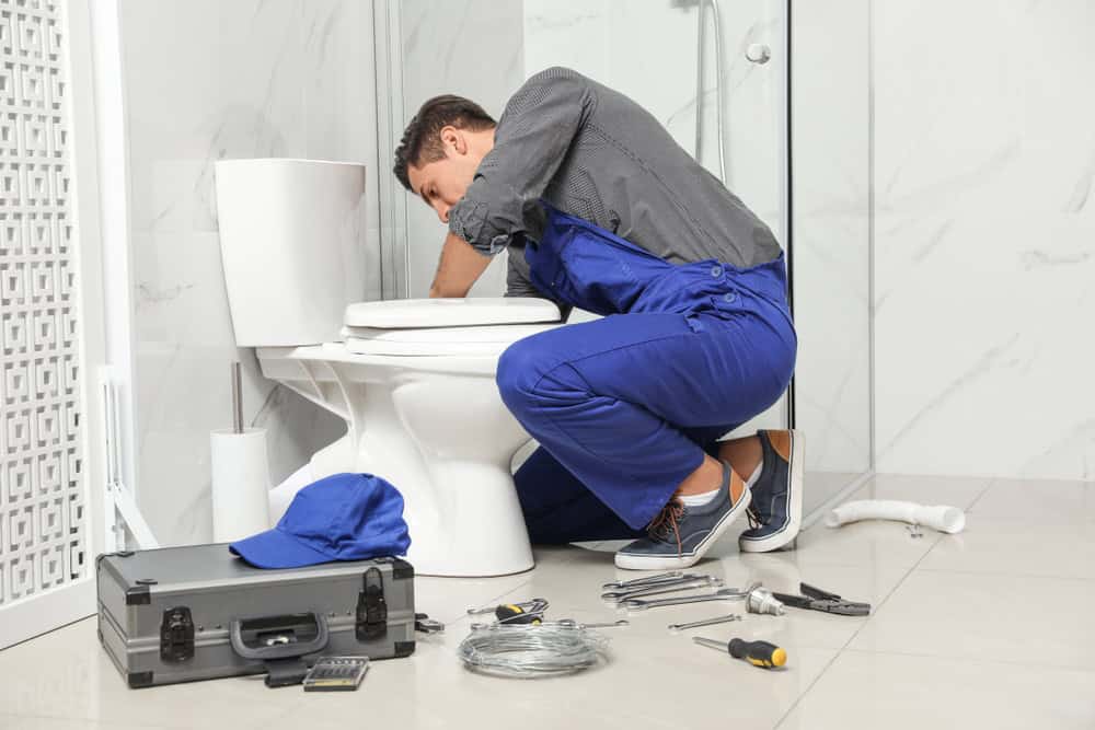 A plumber in blue overalls is kneeling and fixing a toilet in a bathroom. Various tools are scattered on the floor, including wrenches and pliers. A toolbox and a blue cap are nearby.