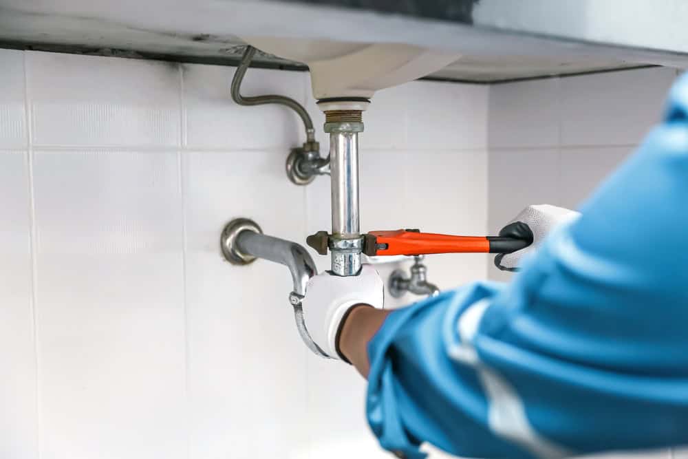 A plumber wearing gloves uses an orange wrench to fix a pipe under a bathroom sink. The scene shows a close-up of the plumber's hands and tools against a white tiled wall.