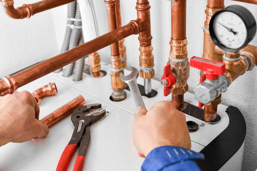 Close-up of a person using a wrench to tighten copper pipes connected to a boiler system. Various pipes and a pressure gauge are visible. Red-handled pliers rest on a surface nearby.