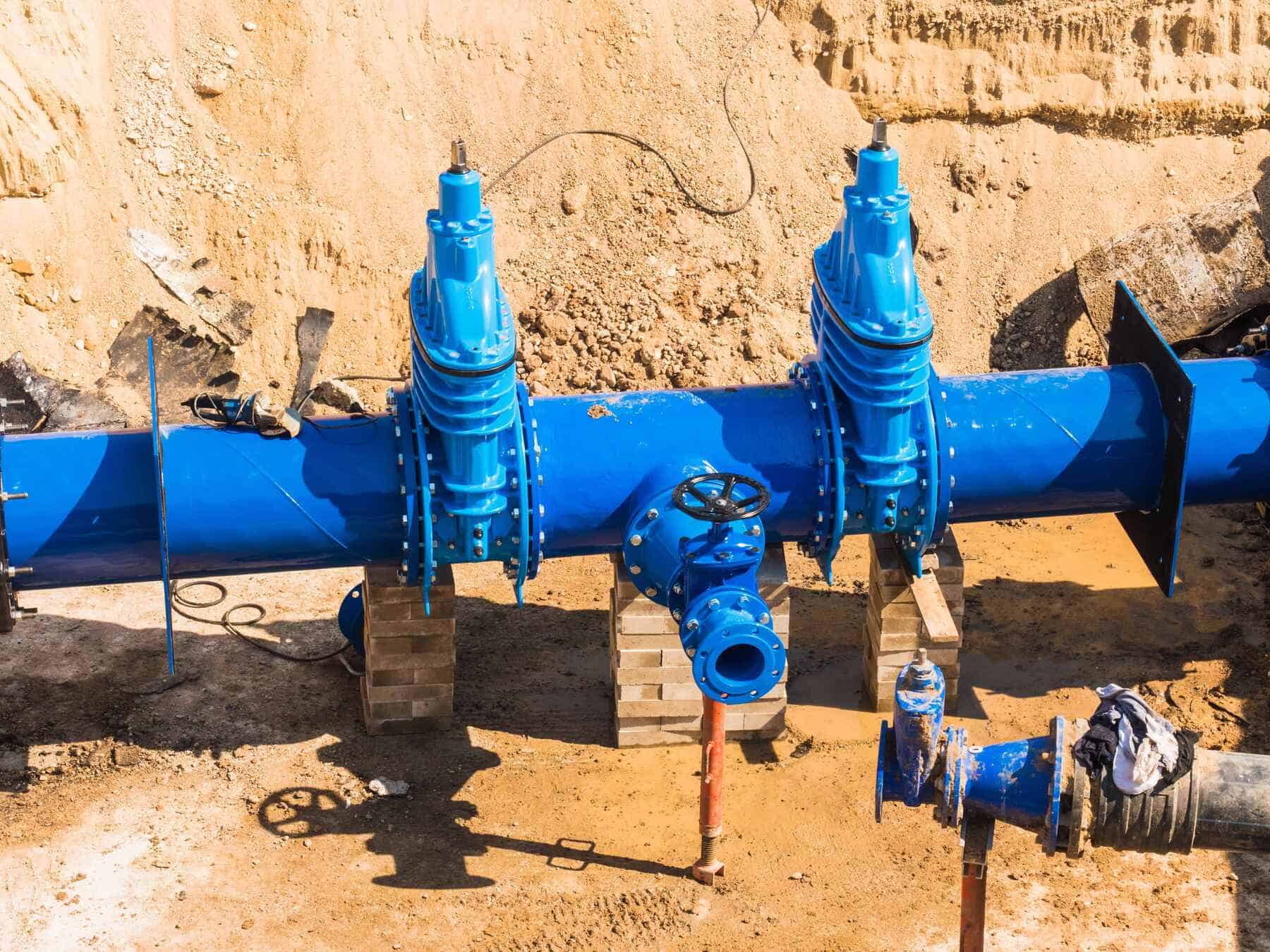 A large blue water pipeline with two valves is installed at a construction site. The pipe is elevated on concrete blocks, surrounded by sand and dirt. Some smaller pipes and construction materials are nearby.