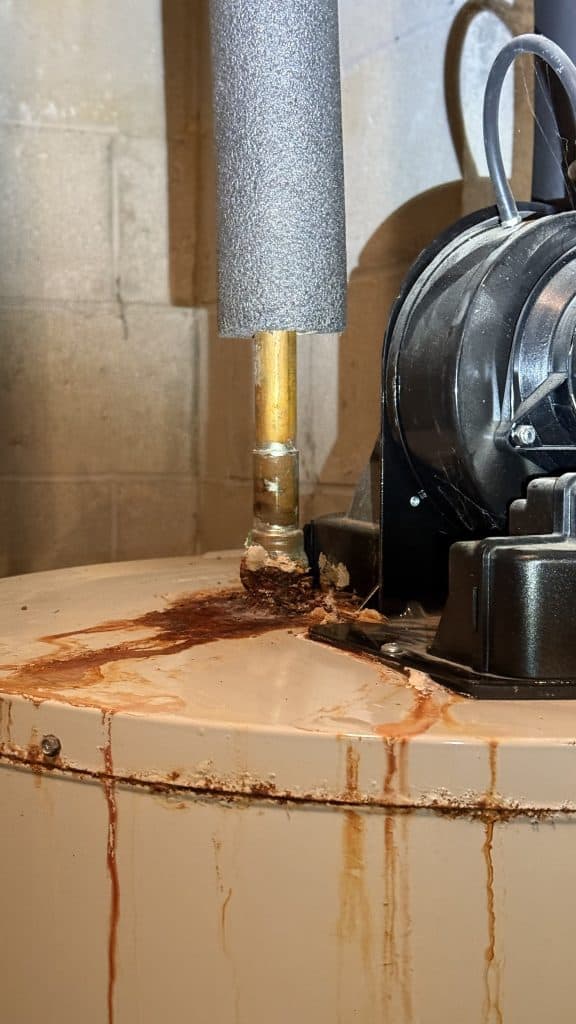 A rusted water heater showing clear signs of leakage. Rust stains and water drips are visible on the surface, along with a pipe insulated with grey foam. The background consists of a grey concrete wall.