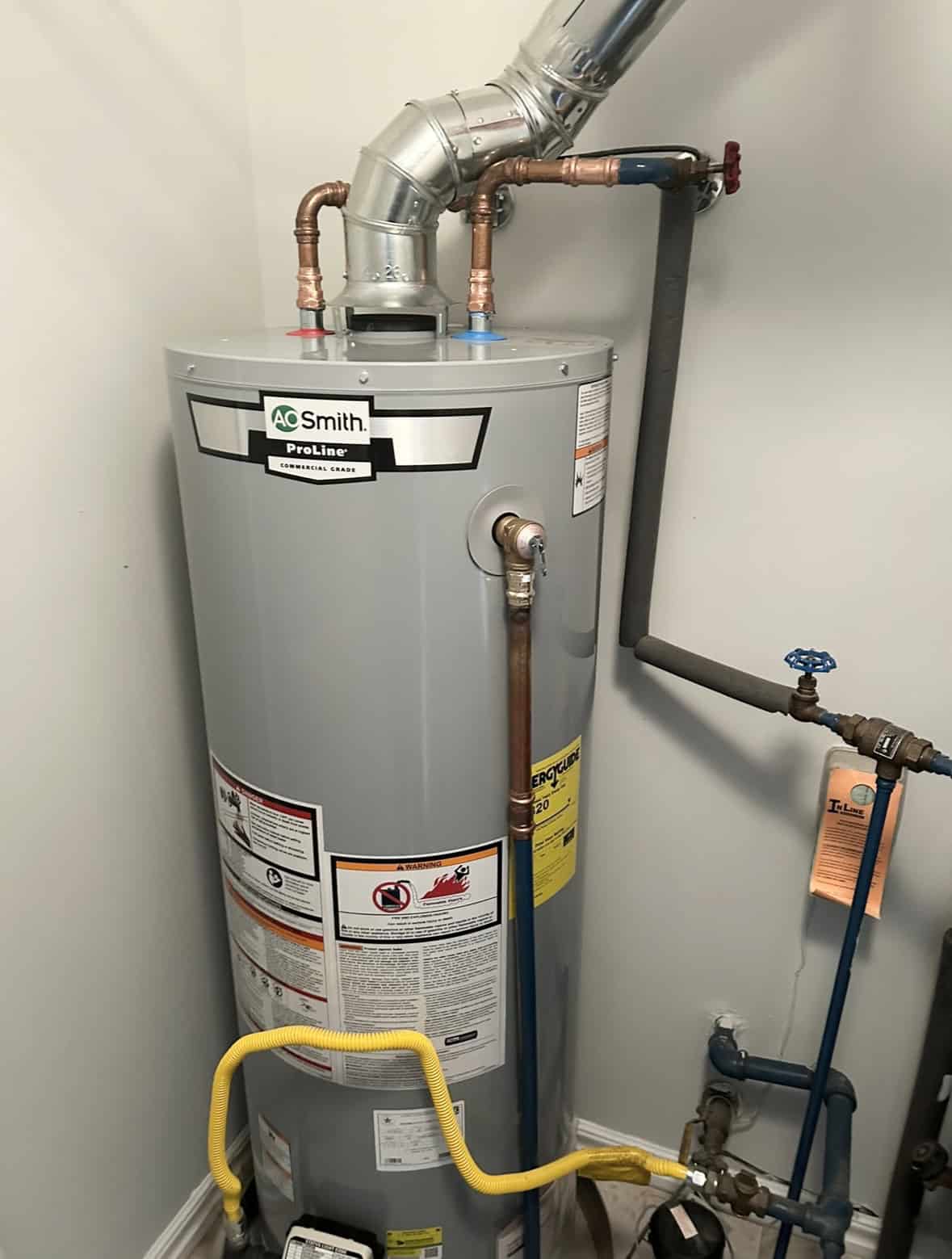 Tall, gray water heater with copper piping, labeled "A.O. Smith ProLine." It is installed in a utility space with gas and water connections. There are warning labels attached, and a blue valve visible on the wall.