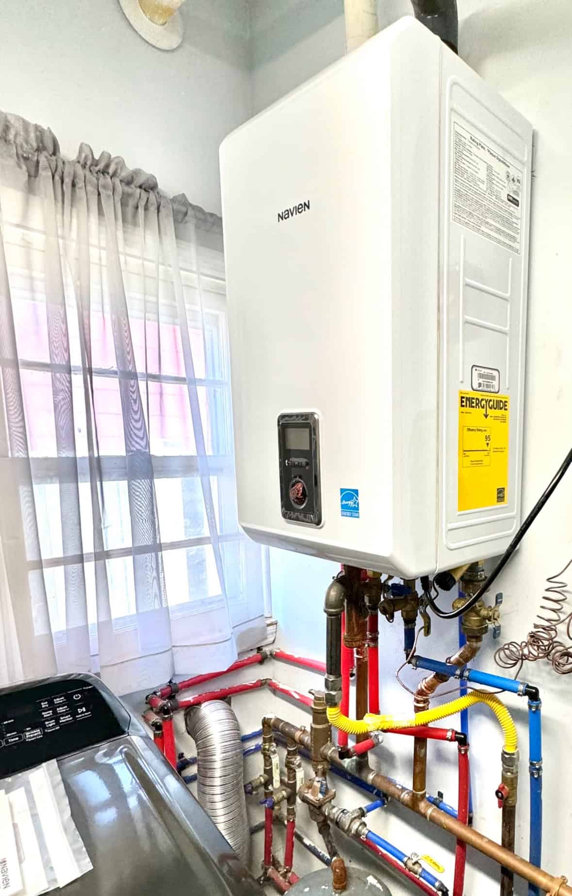 A tankless water heater mounted on a wall next to a window with curtains. Below it are various connected pipes in red, blue, and copper. A yellow energy guide label is attached to the heater. A washing machine is partially visible nearby.