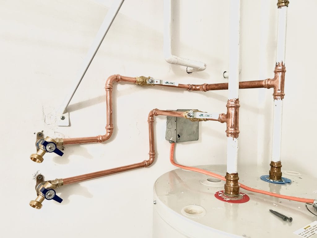 Copper plumbing pipes connected to a water heater tank are shown. The setup includes several joints, blue valve handles, and a white wall background.
