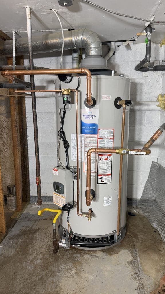 A water heater installed in a basement corner surrounded by pipes. It is a tall, cylindrical unit with copper piping connecting the top, and a ventilation duct extending upwards. The concrete floor and unfinished walls are visible.