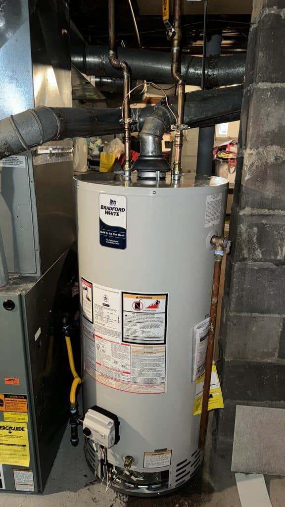 A water heater is installed in a basement, surrounded by pipes and ductwork. It has labels and warnings on its surface. The area is dimly lit and nearby shadows suggest other appliances or items in the vicinity.