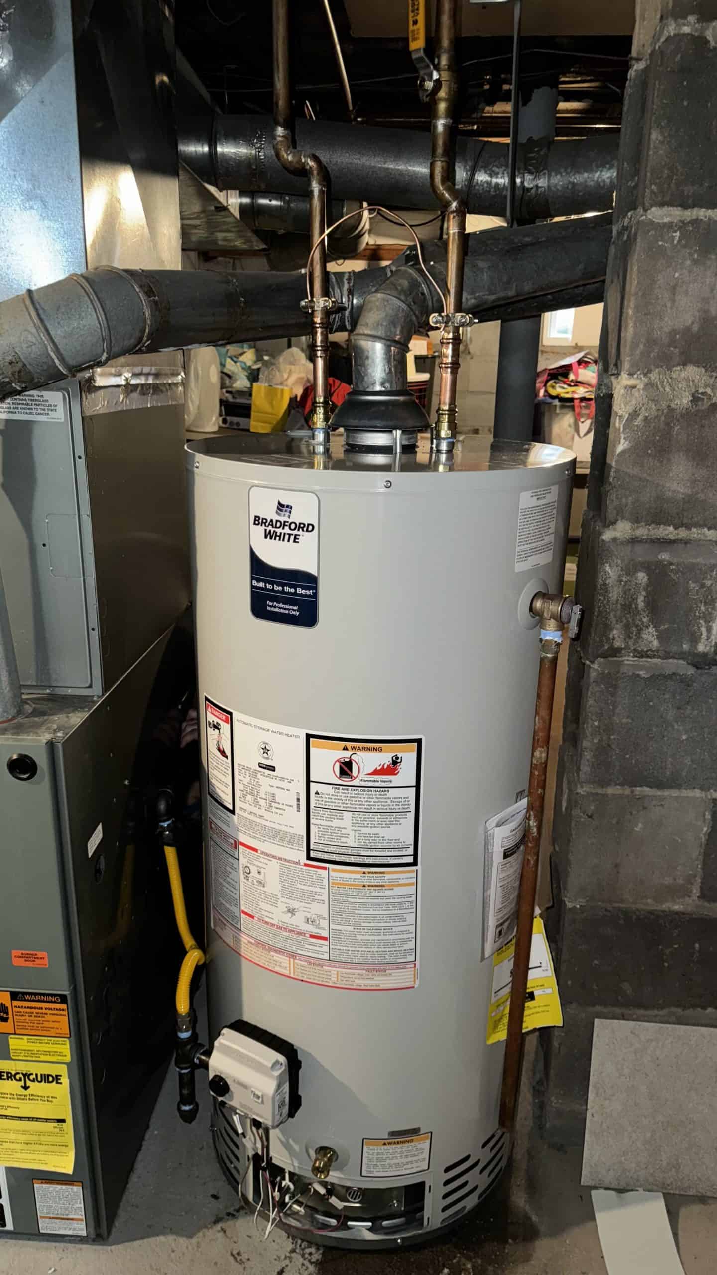 A water heater is installed in a basement, surrounded by pipes and ductwork. It has labels and warnings on its surface. The area is dimly lit and nearby shadows suggest other appliances or items in the vicinity.