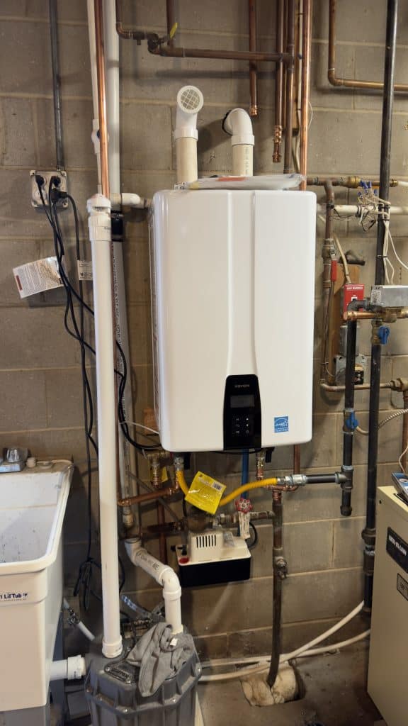 A mounted white tankless water heater with visible pipes and wiring on a concrete wall. Nearby are a utility sink and various plumbing components, including a pressure gauge and valves. A basement setting is suggested.