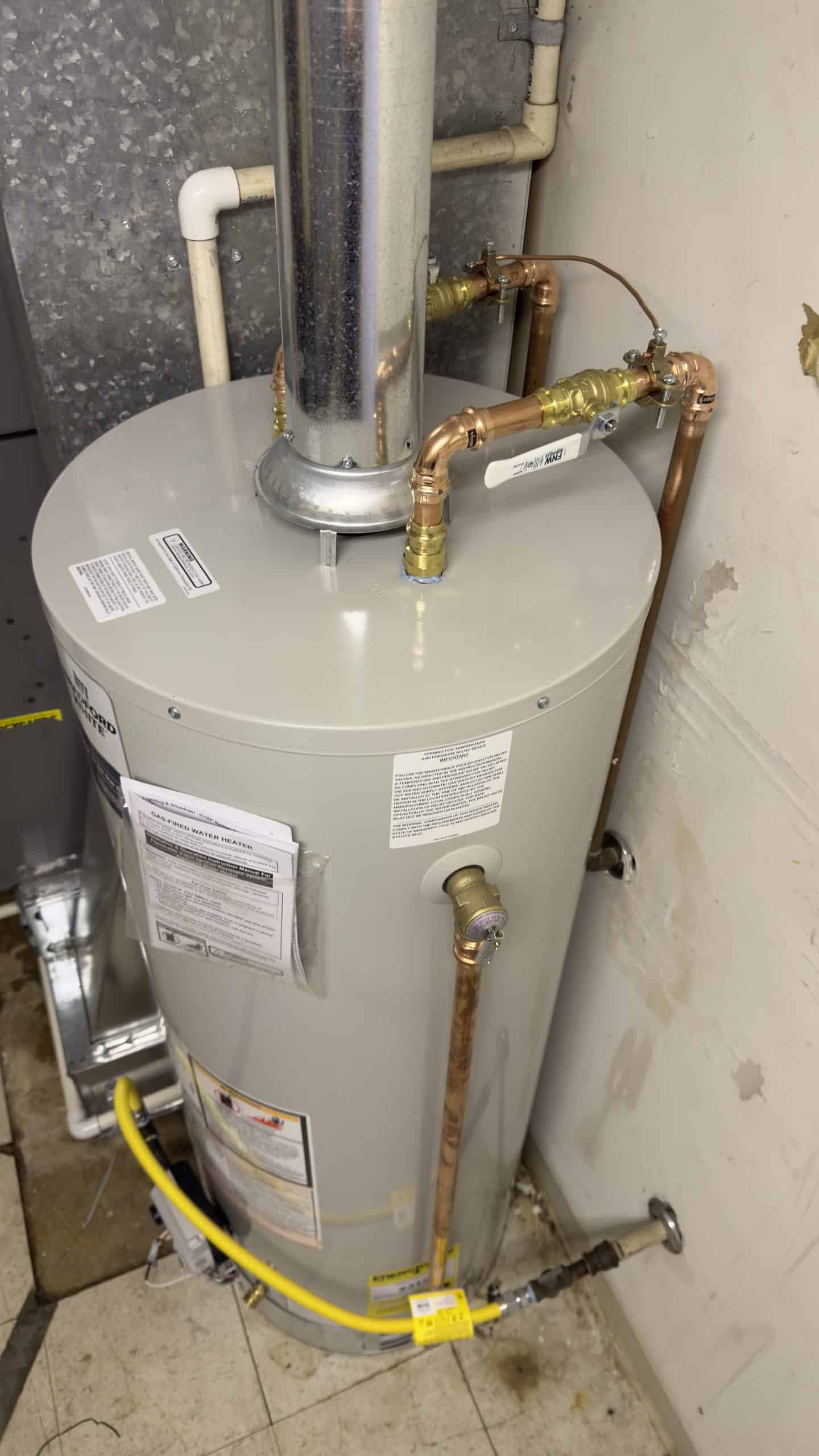 A cylindrical water heater with various copper pipes connected to it is installed in a corner of a room with tiled flooring. There are labels and safety information visible on the tank. A yellow gas line is connected at the bottom.