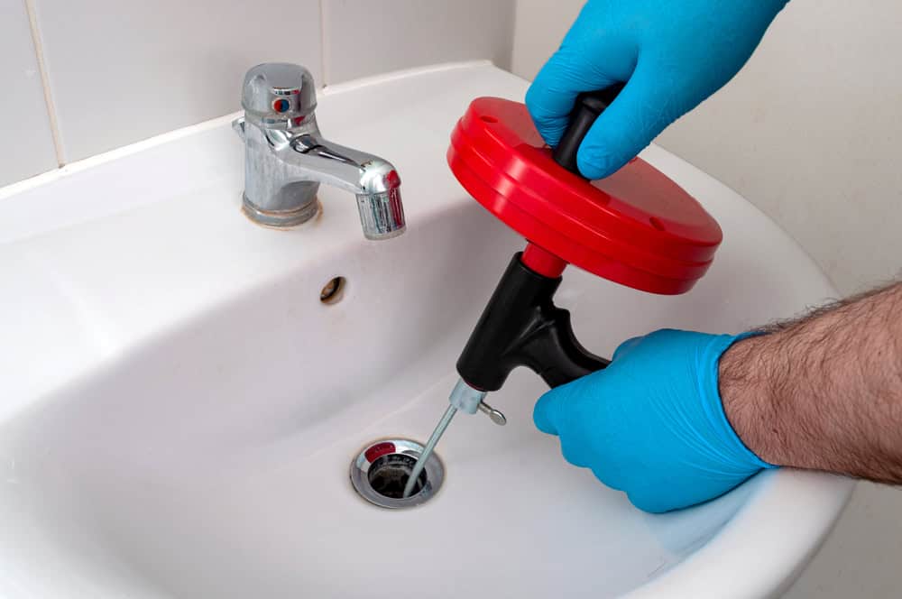 A Plumber in Monmouth County wearing blue gloves uses a red and black drain auger to unclog a white bathroom sink with a silver faucet.