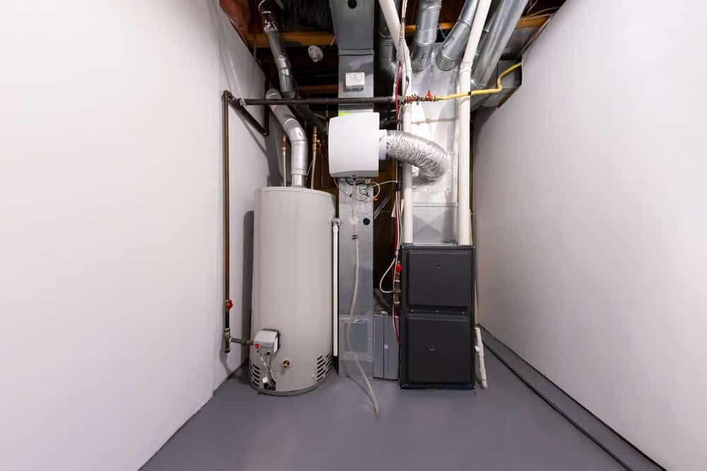 A water heater and furnace with connected ductwork and pipes are expertly installed by a plumber in Monmouth & Ocean County, NJ, in a clean, white-walled basement with a gray floor and exposed ceiling beams.