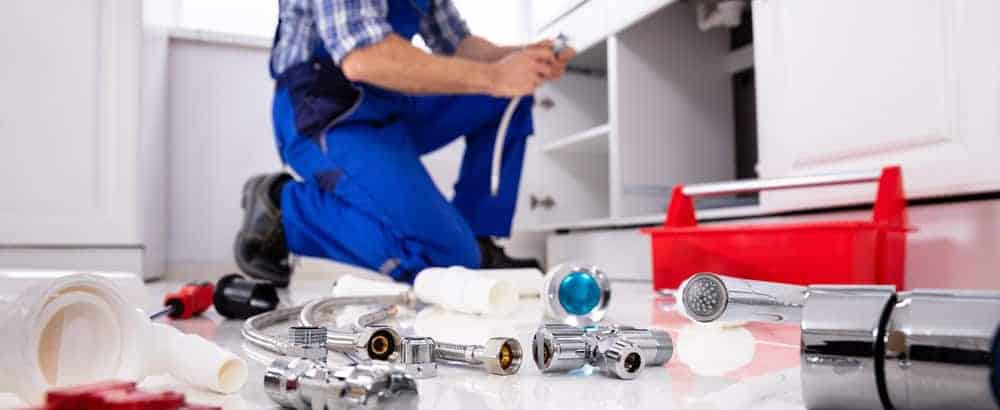 A plumber in Monmouth & Ocean County, NJ, wearing blue overalls, kneels on the floor fixing pipes under a sink, surrounded by plumbing tools and parts spread out on the white floor with a red toolbox nearby.