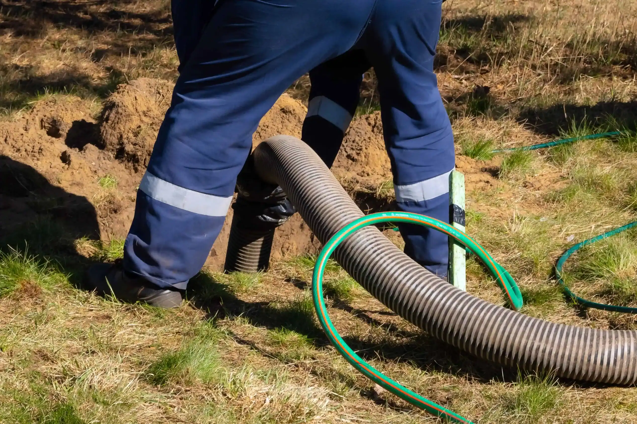 A worker in blue pants and boots operates a large, flexible hose, likely for sewer line cleaning or drainage work, on a grassy area. The ground is partially dug up, and a green hose is also visible on the ground.