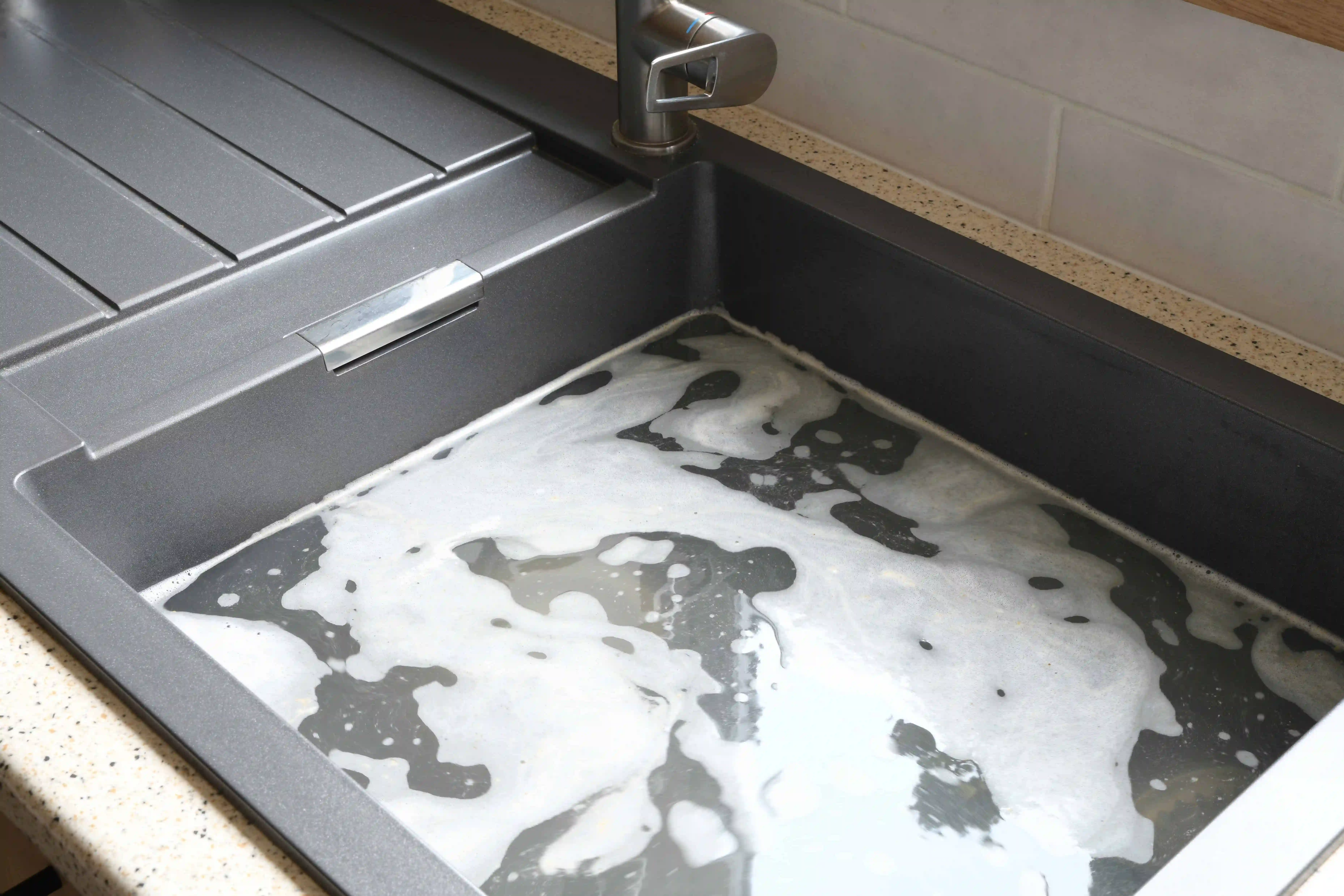 A kitchen sink in NJ filled with soapy water, white soap bubbles floating on the surface. The gray sink and faucet stand out above a beige, speckled countertop—perfect inspiration for any Plumber in Monmouth County.