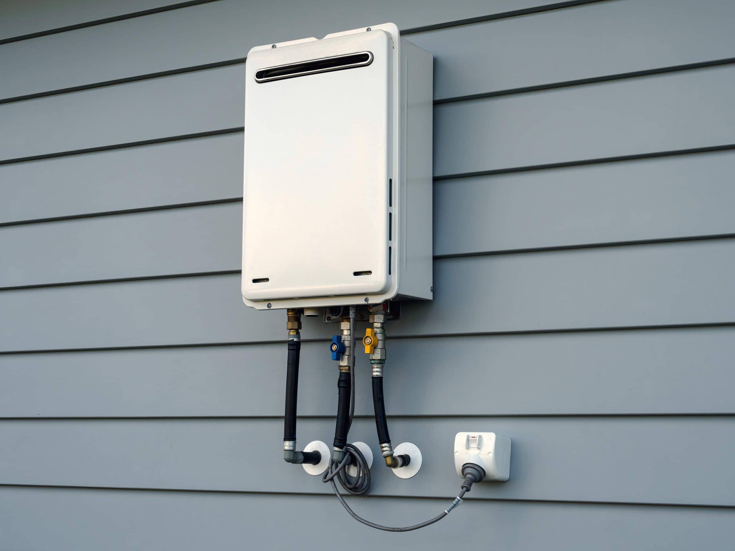 Outdoor tankless water heater mounted on a house exterior with multiple insulated pipes connected below it.