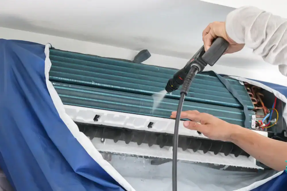 A person uses a high-pressure water spray to clean the interior coils of a wall-mounted air conditioning unit, with a protective cover draped underneath to catch drips.